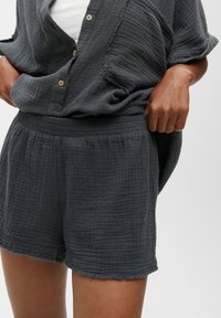 Shorts en tissu tissé léger gris foncé avec une coupe décontractée, taille élastique et ourlet effiloché. Surface texturée avec un motif subtil.