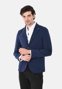 Blazer blu navy in tessuto texturizzato, chiusura a un bottone, revers a punta e due tasche frontali, abbinato a una camicia bianca e pantaloni neri.