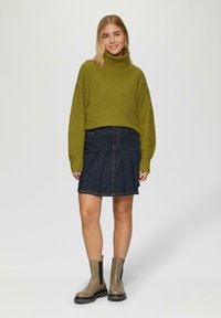 Olijfgroene gebreide trui met hoge kraag, gecombineerd met een donkere denim plissérok en grijze enkellaarsjes met zwarte elastische panelen.