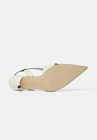 Zapato blanco de punta afilada con un upper de cuero liso, correas negras en los tobillos y suela beige. Presenta un diseño minimalista y acentos redondeados.