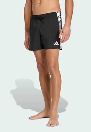 Muž nosící černé plavky Adidas s bílými pruhy po stranách a logem, stojící naboso na světlém pozadí.