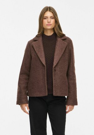 Jeune femme portant un manteau marron texturé par-dessus un pull foncé et un pantalon noir, debout devant un fond clair uni.