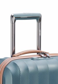 Valigia rigida di color teal testurizzato con manico marrone, doppie cerniere e manico telescopico in metallo. Logo del marchio visibile sul manico.