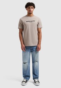 Beige katoenen t-shirt met "BALLIN'" in het zwart bedrukt, gecombineerd met lichtblauwe distressed jeans en zwart-witte sneakers.