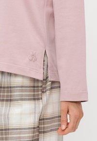 Hand som vilar bredvid en ljusrosa långärmad topp med sidoklyfta och liten upphöjd logotyp, ihop med beige och brun rutiga byxor.