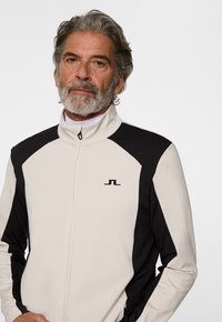 Giacca zip nera e bianca con alto colletto, tessuto texturizzato e logo sul petto. Presenta accenti neri sulle spalle e sulle maniche.