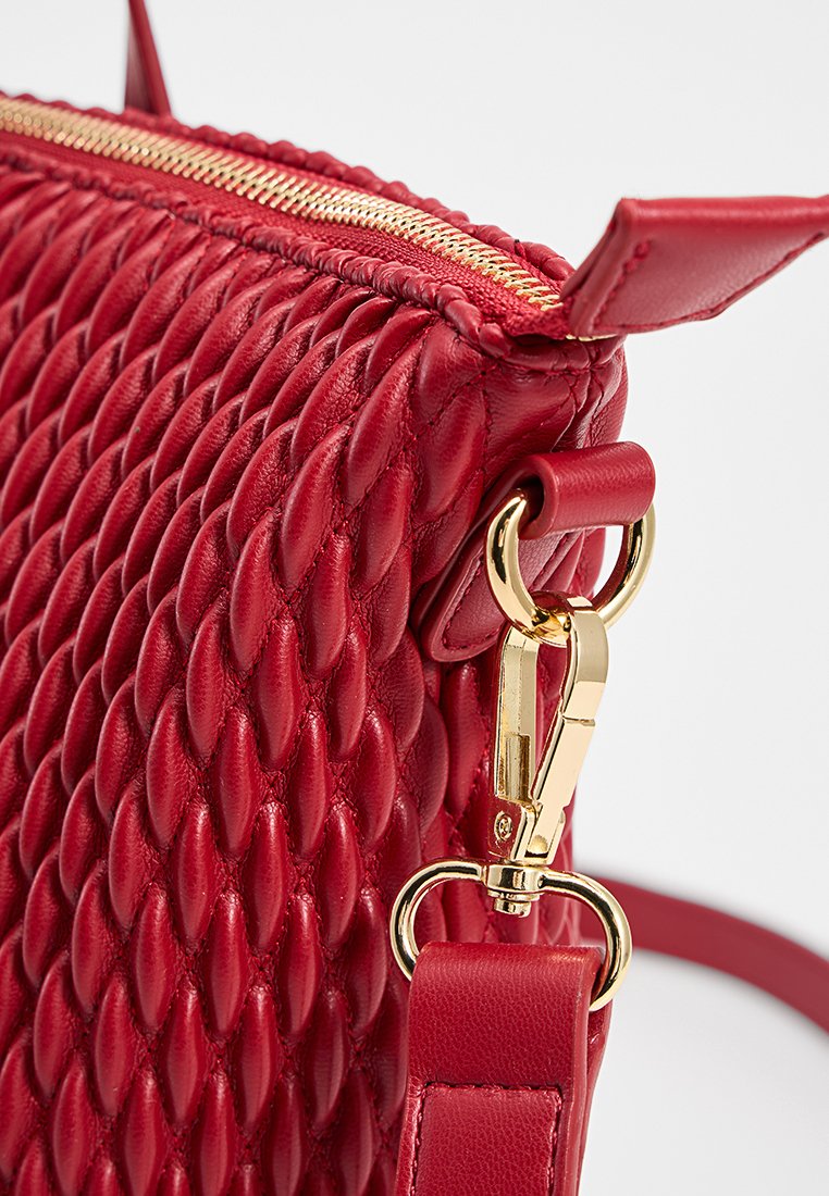 Sac en cuir rouge à bandoulière avec un motif texturé tissé. Comprend une fermeture éclair et des accessoires en métal doré, une bandoulière réglable et une base rigide.