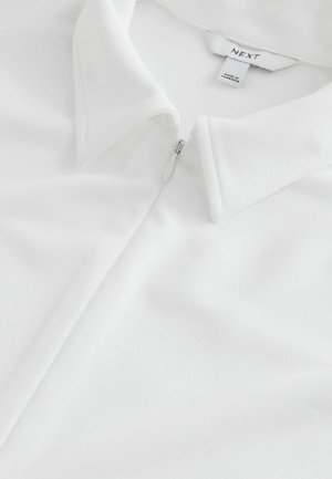Tricou polo alb cu închidere cu fermoar, având o textură moale și design clasic al gulerului. Eticheta brandului "NEXT" este vizibilă în interiorul gulerului.