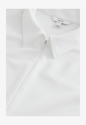 Hvid poloshirt med lynlåslukning, der har en blød tekstur og klassisk kravedesign. Brandlabel "NEXT" synlig indeni kraven.