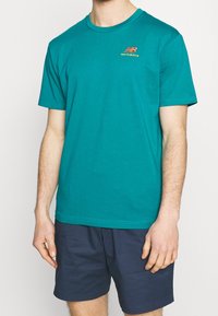 Tealová bavlněná tričko s krátkým rukávem a kulatým výstřihem. Na hrudi se nachází malé vícobarvené logo "New Balance".