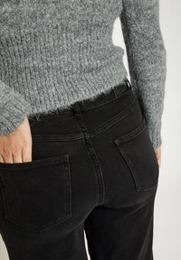 Crne traperice s dva stražnja džepa, uparene s laganim sivim džemperom rebraste pletenine. Fokus na detaljima pojasa i stražnjih džepova.