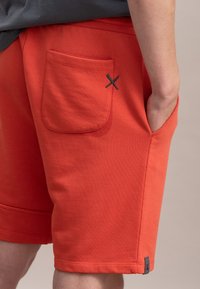 Pantaloni corti rossi in misto cotone, con una tasca laterale con un'accenno contrastante grigio scuro e una finitura liscia e testurizzata.