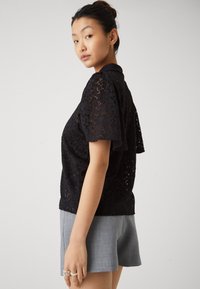 Svart blondebluse med korte pufferm, med blomsterdesign og krage. Kombinert med grå skreddersydde shorts.