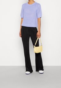 Person, die ein lavendelfarbenes gestricktes Kurzarmoberteil, schwarze plissierte weite Hosen und weiße Sneakers trägt und eine kleine gelbe Handtasche hält.