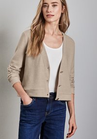 Beige kofta i mjukt tyg med långa ärmar, knappar fram, och rundad halsringning, kombinerad med en vit topp och mörkblå jeans.