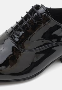 Chaussure en cuir verni noir avec une surface lisse et brillante, un design traditionnel à lacets, et un bout arrondi avec des détails de couture subtils.