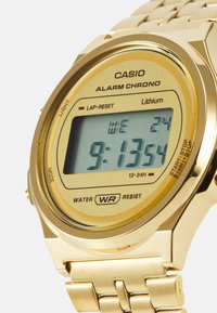 Montre digitale dorée avec un cadran rectangulaire, écran LCD et bracelet en métal. Les caractéristiques incluent l'heure, la date et un indicateur de résistance à l'eau.