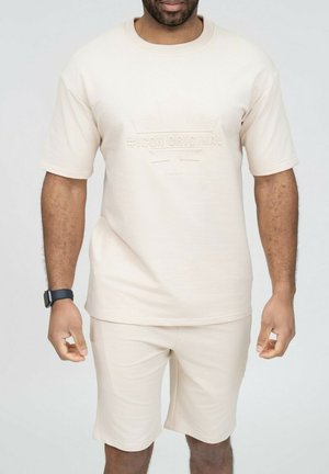 Homme portant une chemise beige à manches courtes et un short assorti avec le texte embossé "ICON ORIGINAL" sur la chemise, debout devant un fond uni.