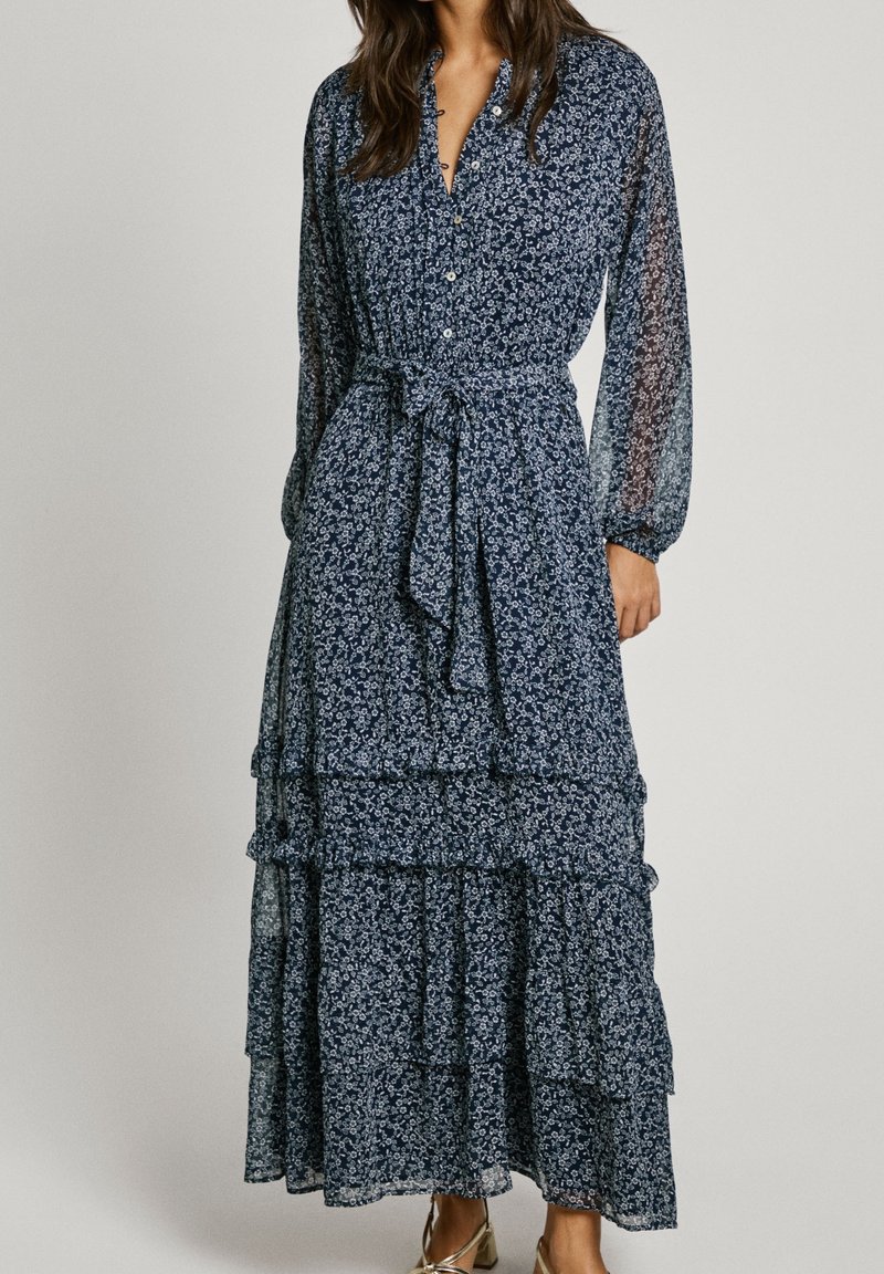 Femme portant une longue robe fleurie bleu marine avec des boutons, une ceinture nouée à la taille, des manches longues transparentes et des couches de jupe à volants.