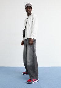 Vit långärmad skjorta, mörkgrå säckiga jeans, röda och marinblå sneakers, och en grå keps, med en liten svart axelväska.