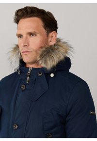 Parka azul marino con capucha forrada de pelo sintético, con botones delanteros, cierre de cremallera y un bolsillo en el pecho para almacenamiento.