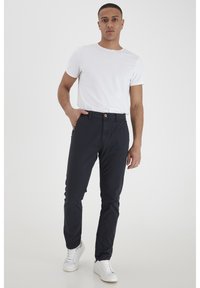 Crne slim-fit hlače od pamuka, s kopčanjem na gumb i džepovima, u kombinaciji s običnom bijelom majicom s okruglim izrezom i bijelim tenisicama.