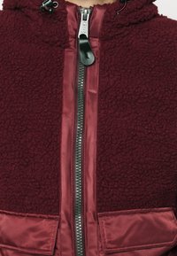 Burgundská fleecová bunda s hladkým saténovým zipem, má vysoký límec, kovový zip a dvě přední kapsy se saténovým povrchem.