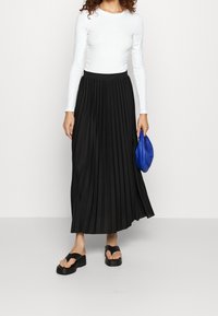 Gonna maxi plissettata nera con vita alta, abbinata a un top aderente bianco a maniche lunghe e sandali con platform neri. Tiene in mano una pochette blu.