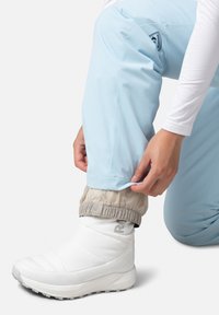 Ljust blå skidbyxor med en åtsittande beige mudd, ihop med en vit isolerad känga. Reflekterande logotyp synlig på byxbenet.