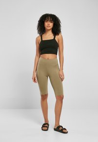 Zwarte crop top met dikke banden, gecombineerd met olijfgroene legging tot midden boven de dij. Het model draagt zwarte instappers met verstelbare banden.