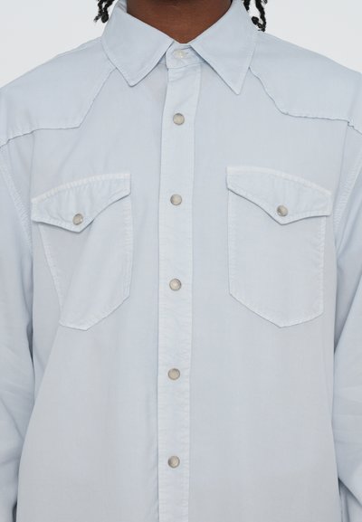 Svetlomodrá košela s gombíkmi vyrobená z mäkkej látky. Má dve vrecká na hrudníku, kovové patentky a klasický golier.