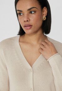 Cárdigan beige con cuello en V y un patrón de punto texturizado, con dos botones y mangas largas. La modelo toca la tela con una mano.