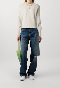 Pull blanc côtelé, jean bleu ample avec ourlet usé, et sac tissé vert dégradé. Le modèle porte des baskets blanches, debout devant un fond neutre.