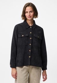 Mlada žena u crnoj kordskoj košulji s dugmadima i dva džepa na prsima te bež hlačama, stoji na jednostavnoj bijeloj pozadini.