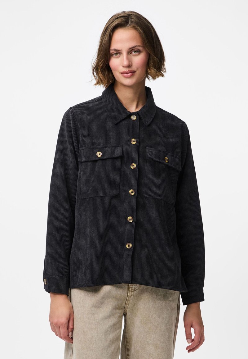 Mlada žena u crnoj kordskoj košulji s dugmadima i dva džepa na prsima te bež hlačama, stoji na jednostavnoj bijeloj pozadini.