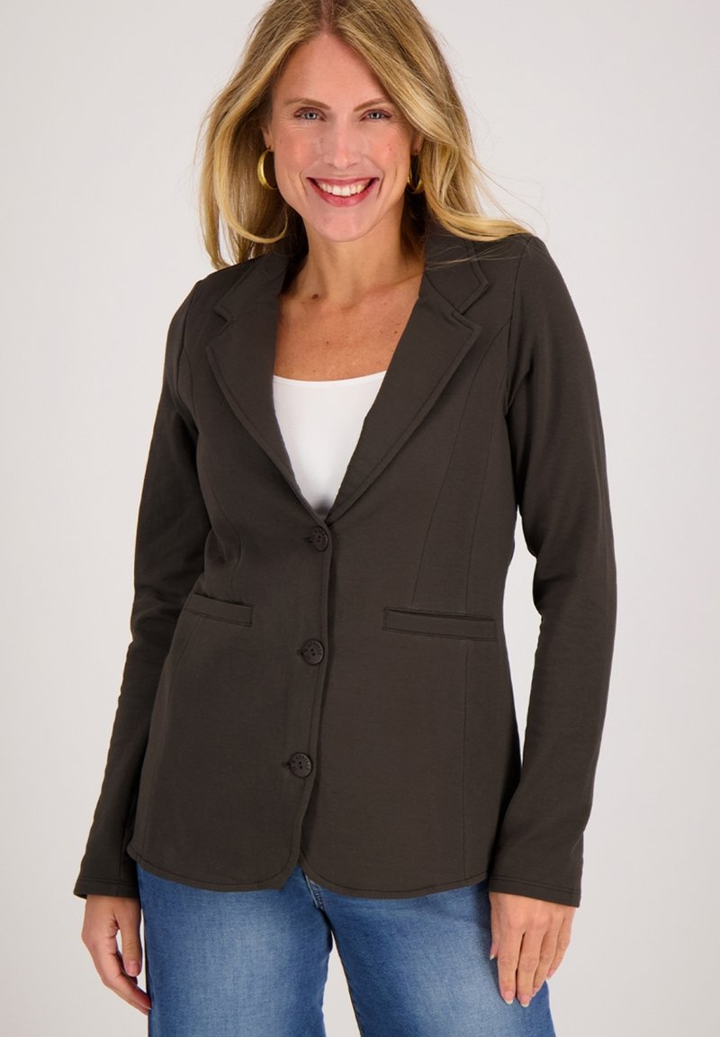 Donkerbruine blazer van zachte stof, met een gelapte kraag, drie knopen en twee voorzakken, gedragen met een witte top en jeans.