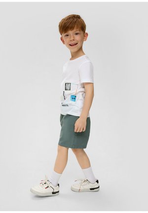 Biała koszulka bawełniana z nadrukami graficznymi, w połączeniu z zielonymi szortami i białymi sneakersami. Stylizacja jest swobodna i wygodna dla dzieci.