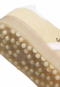 Αθλητικό παπούτσι με υφή κίτρινο πάνω μέρος, λευκή φτέρνα και διαφανή καουτσούκ σόλα με εξαγωνικά μοτίβα πρόσφυσης. Ροζ branding στο πλάι.