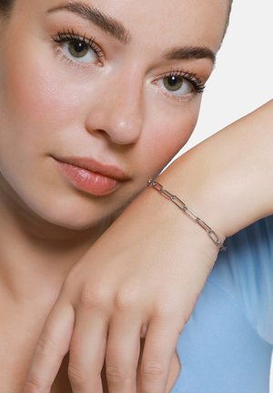 Srebrna bransoletka łańcuszkowa z wydłużonymi ogniwami. Noszona na nadgarstku modelki, w zestawieniu z jasnoniebieską bluzką, podkreślająca elegancki, minimalistyczny design.