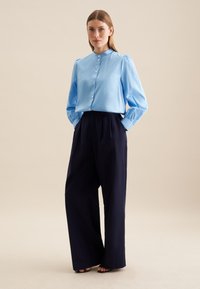 Hellbluse mit hohem Kragen und Knopfleiste. Kombiniert mit navyblauen, hoch taillierten, weiten Hosen. Glattes Material und taillierte Passform.