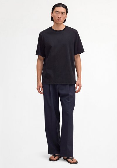 Mann steht vor weißem Hintergrund, trägt ein schwarzes übergroßes T-Shirt, marineblaue, weit geschnittene Hosen und schwarze Sandalen.