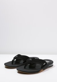 Fekete flip-flop papucs, textúrált gumi felsőrésszel, párnázott talppal és barna gumitalppal; a pántján logó részlet található.