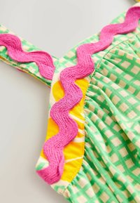 Tissu à carreaux vert et blanc avec des bordures en forme de coquille rose et des accents jaunes. Présente une texture douce et des bretelles ajustables.