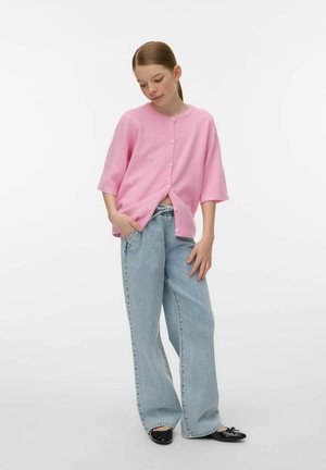 Jeune personne portant un cardigan rose ample boutonné, un jean large bleu clair et des ballerines noires, debout les mains dans les poches.