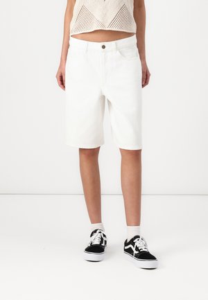 Shorts en denim blanc avec une coupe droite, dotés d'une fermeture à bouton à l'avant et de cinq poches. Associés à des baskets noires et blanches.