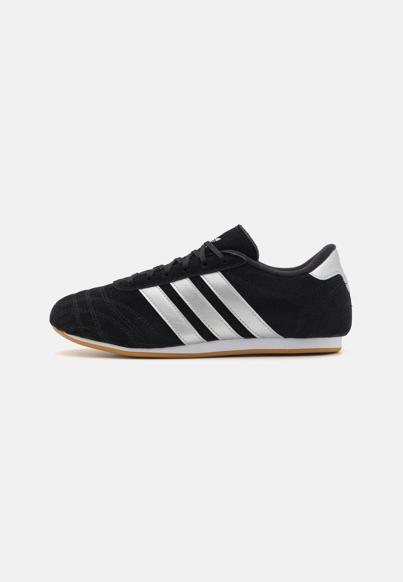 Black suede sneaker with three silver stripes, rubber sole, rounded toe, and textured upper for grip. Angular design, sporty finish.