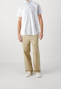 Hvid kortærmet skjorte med knaplukning og subtil tekstur, parret med beige løstsiddende bukser og hvide sneakers. Enkel, afslappet design.