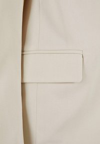 Tessuto di blazer beige con una texture liscia e una tasca a patta. Presenta linee pulite con dettagli di cucitura visibili.