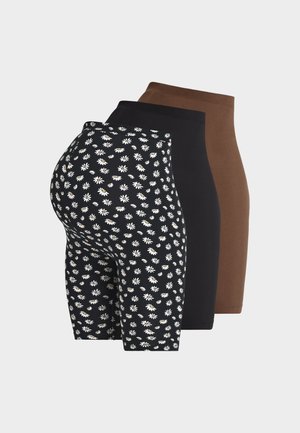 Femme enceinte debout portant un t-shirt noir, un short à fleurs noir et blanc, des ballerines rouges avec des liens à la cheville, tenant un grand sac cabas noir.
