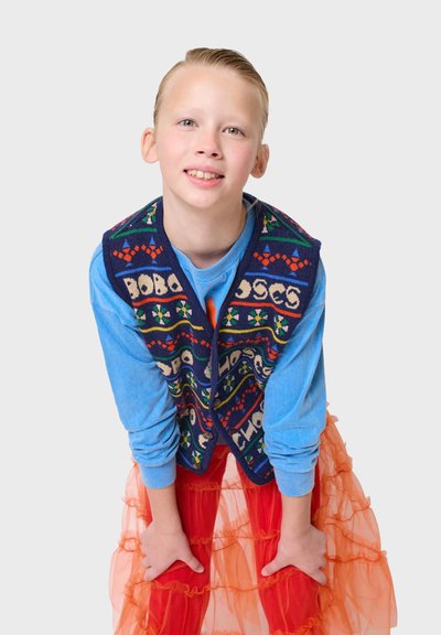 Blå långärmad skjorta under en färgglad stickad väst med mönster i blått, rött och grönt, kombinerad med en lager av orange tyllkjol.